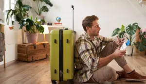 Smiling handsome guy looks at his passport and flight ticket, sits on floor with suitcase.