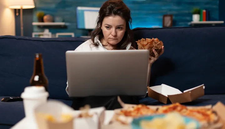 Caucasian female sitting on sofa eating tasty delicious burger while working on laptop computer in living room late at night. Woman enjoying takeaway food home delivered. Fastfood lunch meal order