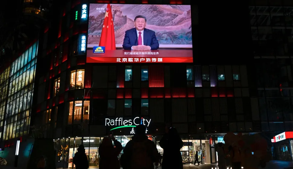 People stand beneath a large screen showing China's President Xi Jinping delivering a speech ahead of the New Year's eve celebrations in Beijing on December 31, 2025. China's President Xi Jinping said on December 31 that "reunification...is unstoppable" as he addressed the nation shortly after Beijing's military announced the end of live-fire drills around Taiwan. (Photo by WANG Zhao/AFP)