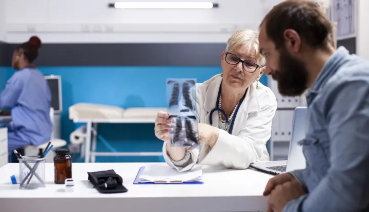 In clinic, elderly female health specialist holds xray, pointing out diagnosis details for young man with beard. Senior doctor advices male patient regarding radiology results and medical next steps.rak pluća doktor liječnik pacijent ilustracija freepik