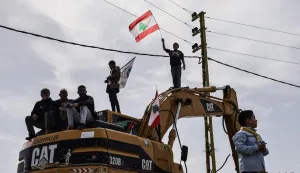 Standing on a digger used to clear the rubble of destroyed homes abd businesses, a Lebanese youth holds up the Lebanese flag as residents wait for the arrival of Lebanon's prime minister during his visit to the heavily-damaged southern village of Kfar Shouba, near the border with Israel on February 8, 2026. Swathes of south Lebanon's border areas remain in ruins and largely deserted more than a year after a US-brokered November 2024 ceasefire sought to end hostilities between Israel and the Iran-backed group. Salam vowed authorities would begin key projects including restoring roads, communications networks and water in the two towns (Photo by Rabih DAHER/AFP)