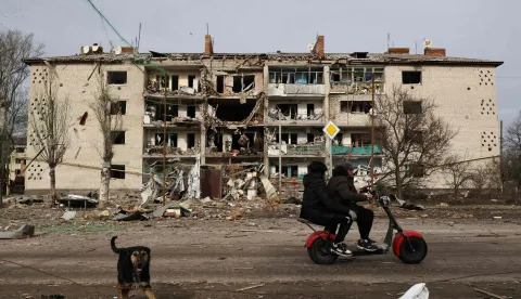 TOPSHOT - This handout photograph taken and released by the press service of the 65th Mechanized Brigade of the Ukrainian Armed Forces on February 20, 2026, shows a dog and local residents riding an electric bike past a damaged residential building following an alleged air attack in Komyshuvakha, Zaporizhzhia region, amid the Russian invasion of Ukraine. (Photo by Andriy Andriyenko/65th Mechanized Brigade of Ukrainian Armed Forces/AFP)/RESTRICTED TO EDITORIAL USE - MANDATORY CREDIT "AFP PHOTO/65TH MECANIZED BRIGADE OF UKRAINIAN ARMED FORCES" - HANDOUT - NO MARKETING NO ADVERTISING CAMPAIGNS - DISTRIBUTED AS A SERVICE TO CLIENTS - AFP CANNOT INDEPENDENTLY VERIFY THE AUTHENTICITY OR LOCATION, DATE, AND CONTENT OF THESE IMAGES./