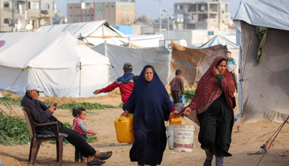 Women walk with pails of water and jerry cans after filling up from a mobile cistern in the Maghazi camp for Palestinian refugees in the central Gaza Strip on February 11, 2026. Since October 10, a fragile US-sponsored truce in Gaza has largely halted the fighting between Israeli forces and Hamas, but both sides have alleged frequent violations. (Photo by Eyad BABA/AFP)