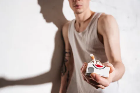 Cropped photo of young attractive man standing on floor with cigarette posing isolated over wall background. Offers cigarettes to camera.