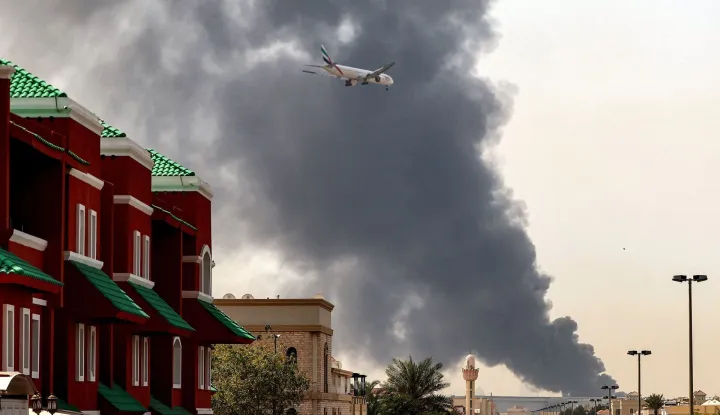 An Emirates aircraft prepares for landing as a smoke plume rises from an ongoing fire near Dubai International Airport in Dubai on March 16, 2026. Flights were gradually resuming at Dubai airport on March 16, previously the world's busiest for international flights, the airport operator said, after a "drone-related incident" sparked a fuel tank fire nearby, as Iran kept up its Gulf attacks. (Photo by AFP)/