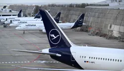 (FILES) The logo of German airline Lufthansa is seen on vertical stabilisers of aircraft parked on the tarmac while in the background an airplace is seen in flight at the international Frankfurt Airport, Frankfurt am Main, western Germany, on March 12, 2026. Cabin crew at German airline Lufthansa will go on strike on April 10, 2026 over an ongoing labour dispute, their union said. (Photo by Kirill KUDRYAVTSEV/AFP)