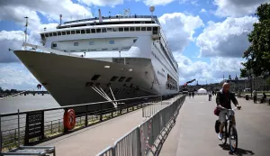 A man rides his bicycle past the Bahamas-registered cruise ship Ambition after passengers were confined following the outbreak of a gastrointestinal illness onboard, at the Bordeaux port in Bordeaux, southwestern France on May 13, 2026. French authorities on May 13 confined more than 1,700 passengers and crew on a British cruise ship docked in Bordeaux after an elderly passenger died, said officials, who played down any links to the hantavirus scare. (Photo by Christophe ARCHAMBAULT/AFP)
