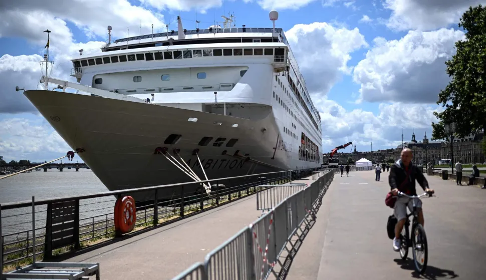 A man rides his bicycle past the Bahamas-registered cruise ship Ambition after passengers were confined following the outbreak of a gastrointestinal illness onboard, at the Bordeaux port in Bordeaux, southwestern France on May 13, 2026. French authorities on May 13 confined more than 1,700 passengers and crew on a British cruise ship docked in Bordeaux after an elderly passenger died, said officials, who played down any links to the hantavirus scare. (Photo by Christophe ARCHAMBAULT/AFP)