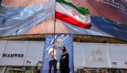 A man raises an Iranian national flag as he stands on a podium before a political billboard featuring US President Donald Trump and the Strait of Hormuz along Valiasr Square in Tehran on May 26, 2026. US forces launched overnight strikes on missile sites in Iran and boats they said were trying to lay mines in Gulf waters, sending oil prices higher on May 26, while diplomats sought a deal to end the war. Iran did not immediately confirm the reported strikes, but state media reported blasts in the southern port city of Bandar Abbas, and the Revolutionary Guards Corps said its forces had downed a US drone entering its airspace and fired at an F-35 fighter jet. (Photo by ATTA KENARE/AFP)