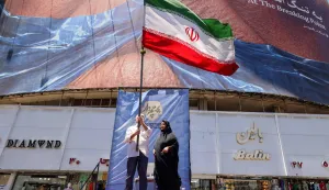 A man raises an Iranian national flag as he stands on a podium before a political billboard featuring US President Donald Trump and the Strait of Hormuz along Valiasr Square in Tehran on May 26, 2026. US forces launched overnight strikes on missile sites in Iran and boats they said were trying to lay mines in Gulf waters, sending oil prices higher on May 26, while diplomats sought a deal to end the war. Iran did not immediately confirm the reported strikes, but state media reported blasts in the southern port city of Bandar Abbas, and the Revolutionary Guards Corps said its forces had downed a US drone entering its airspace and fired at an F-35 fighter jet. (Photo by ATTA KENARE/AFP)