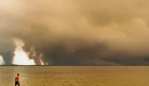 CORRECTION/TOPSHOT - A beachgoer walks in the lake Lacanau, also called Etang de Lacanau (Lacanau Pond) from the beach of Moutchic in Lacanau, southwestern France on July 24, 2026, as clouds of smoke rise into the sky from a wildfire. The fire raging in the northern part of the Arcachon Basin in Gironde, southwest France, has swept through more than 3,400 hectares, according to the latest figures released by the prefecture. More than 20,000 residents and tourists have been evacuated from this part already devastated by flames in 2022. Scientists have long warned that climate change caused by human use of fossil fuels will make periods of drought more intense and longer-lasting, creating the ideal conditions for wildfires. (Photo by Maximilien LAMY/AFP)/?The erroneous mention[s] appearing in the metadata of this photo by Maximilien LAMY has been modified in AFP systems in the following manner: [lake Lacanau] instead of [the ocean]. Please immediately remove the erroneous mention[s] from all your online services and delete it (them) from your servers. If you have been authorized by AFP to distribute it (them) to third parties, please ensure that the same actions are carried out by them. Failure to promptly comply with these instructions will entail liability on your part for any continued or post notification usage. Therefore we thank you very much for all your attention and prompt action. We are sorry for the inconvenience this notification may cause and remain at your disposal for any further information you may require.?