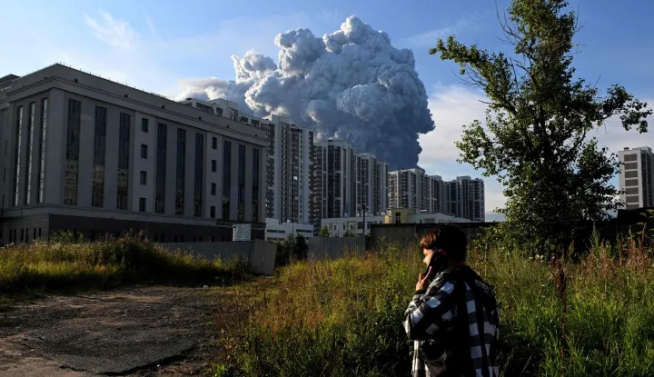 A person speaks on a mobile phone as smoke rises above residential buildings from a warehouse for a Russian e-commerce giant near Saint Petersburg after being hit by Ukrainian drones early on July 24, 2026 morning, triggering a fire that sent smoke pouring over the historic city, according to local officials. Kyiv has targeted Saint Petersburg as it steps up long-range drone strikes on Russian energy, military and logistics sites in recent weeks. Russia's defence ministry said its forces shot down some 571 drones overnight over more than a dozen regions and Crimea, the Black Sea peninsula that Russia annexed in 2014. (Photo by AFP)