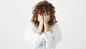 Studio portrait of tired or bothered woman hiding face in her curly hair and holding hands on it, expressing exhaustion or just being childish, standing over gray background. I am not seeing you.