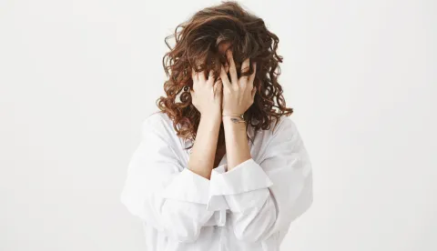 Studio portrait of tired or bothered woman hiding face in her curly hair and holding hands on it, expressing exhaustion or just being childish, standing over gray background. I am not seeing you.