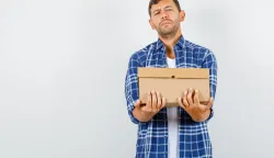 Young man holding heavy cardboard box in shirt, front view. kutija paket online shopping narudzba 