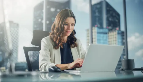 Portrait of Successful Young Businesswoman Sitting at Her Desk Working on Laptop Computer in Big City Office. Confident Professional CEO Managing Environmental, Social and Corporate Governance