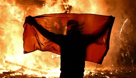 TOPSHOT - A protester holds an Albanian National flag in front of a burning kiosk during an anti-government demonstration in central Tirana on October 15, 2026, as protests continued for a third consecutive day over university conditions and demands for the resignation of Albanian Prime Minister. The latest rally follows two nights of violence in the capital, during which protesters and riot police clashed, police used water cannons and tear gas, and demonstrators damaged government offices and shops and set fire to several buildings. Armend NIMANI/AFP (Photo by Armend NIMANI/AFP)