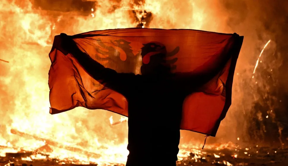 TOPSHOT - A protester holds an Albanian National flag in front of a burning kiosk during an anti-government demonstration in central Tirana on October 15, 2026, as protests continued for a third consecutive day over university conditions and demands for the resignation of Albanian Prime Minister. The latest rally follows two nights of violence in the capital, during which protesters and riot police clashed, police used water cannons and tear gas, and demonstrators damaged government offices and shops and set fire to several buildings. Armend NIMANI/AFP (Photo by Armend NIMANI/AFP)