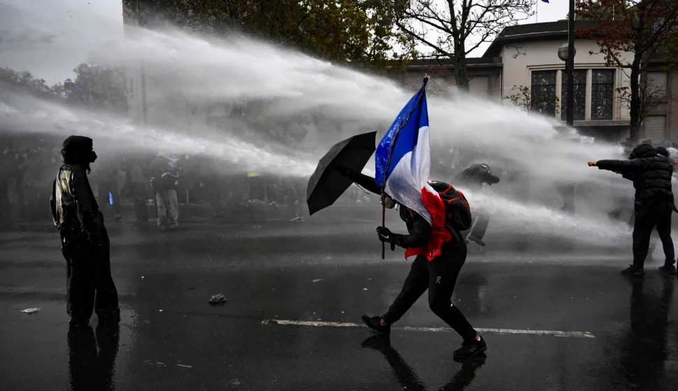 TOPSHOT - A protester holds the French flag as he protects himself from a water cannon during a demonstration in favor of purchasing power, the protection of public services, and fiscal and social justice, in Paris on October 24, 2026 as part of a nationwide call for mobilisation bringing together former members of the Yellow Vest movement, labor unions, high school and university students. Protests by high school students over learning conditions sparked a movement that has since spread nationwide, fuelled by broader anger over the rising cost of living. About 70,000 security personnel will be deployed on October 24, 2026, Interior Minister said at a news conference, warning that calls for violence and looting were circulating on social media. (Photo by Aris MESSINIS/AFP)