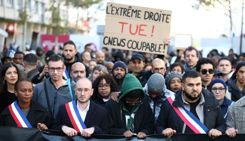 TOPSHOT - Attendees and officials including mayor of Aubervilliers Sofienne Karroumi (R) with an attendee in the background holding a placard reading "the far right kills ! CNEWS guilty !" referencing French TV channel, take part in a silent march in tribute to Pengo Bossio Wangi, a man killed in a shooting by an attacker on October 23, a march organised by his MMA club near the town hall of Aubervilliers, north of Paris, on October 25, 2026. A man suspected of a gun attack in the Paris suburbs that killed Pengo Bossio Wangi and wounded five other persons has left custody and been placed in a psychiatric unit for observation, French anti-terror prosecutors said on October 25, 2026. The 39-year-old suspect, partly identified as Kevin T., was arrested on October 23, in possession of a submachine gun and tear-gas grenades in his car, shortly after the victims were shot as a vehicle drove through the working-class town of Trappes and neighbouring La Verriere. (Photo by Sameer AL-DOUMY/AFP)